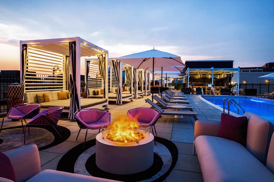 Luxury resort deck at dusk with illuminated cabanas, fire pit, purple lounge chairs, and pool