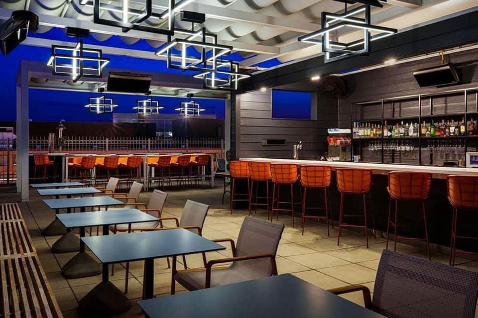 Modern rooftop bar lounge with blue ceiling, orange chairs, black tables, and nighttime water views