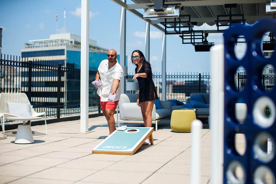 Two guests playing cornhole on rooftop terrace with city skyline and waterfront views