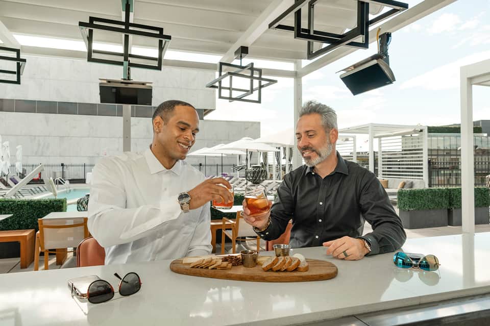 Two men enjoying drinks on modern rooftop bar overlooking pool with contemporary architecture