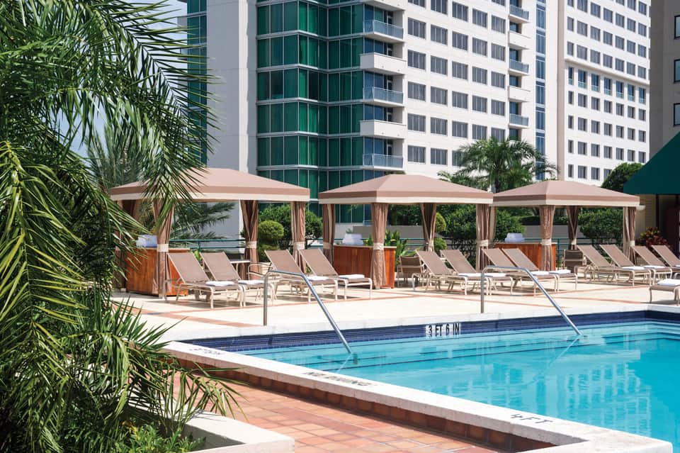Resort pool surrounded by tall apartment buildings with tan umbrellas, lounge chairs, and palm trees