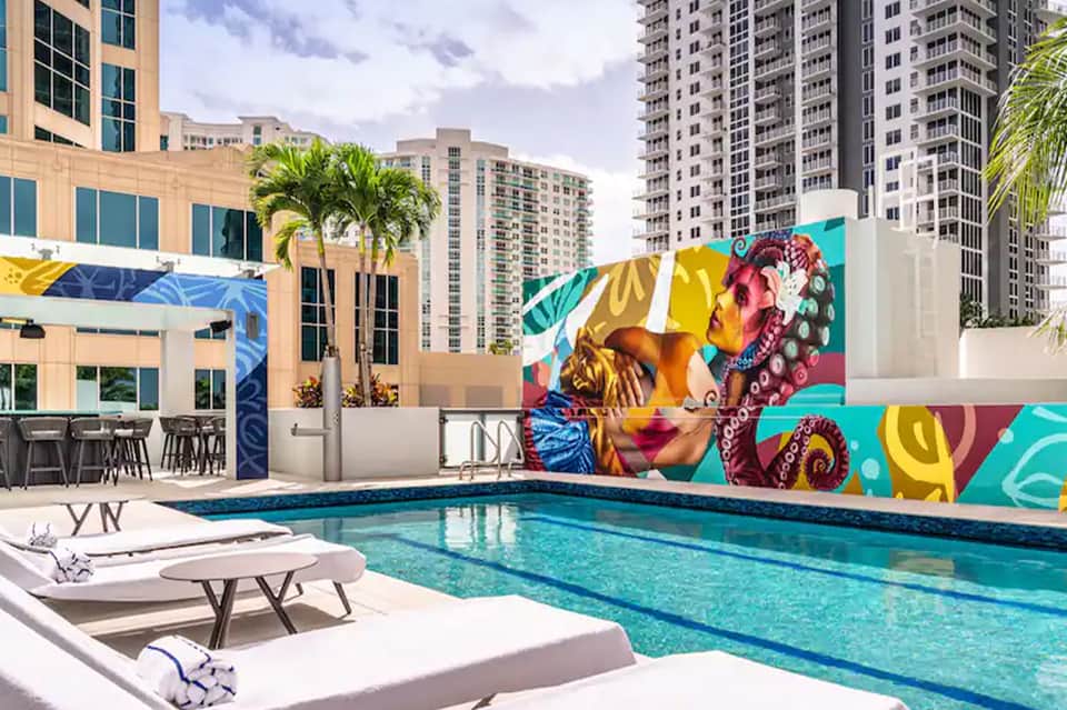 Urban rooftop pool with colorful pop-art mural, lounge chairs, and city buildings