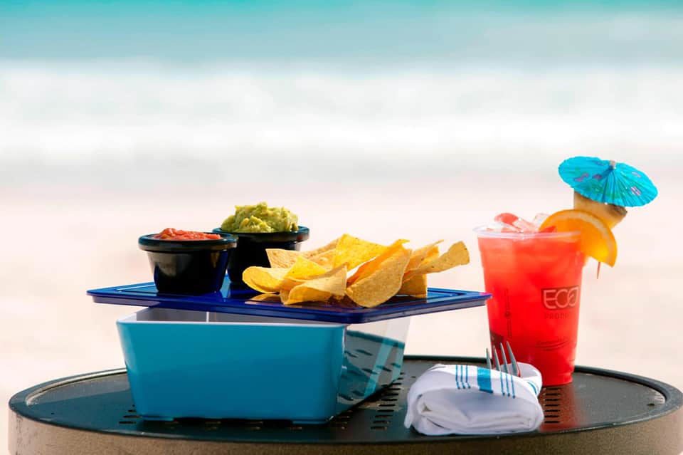 Beach table setting with tropical cocktail, snacks, and decorative umbrella against ocean and sky backdrop