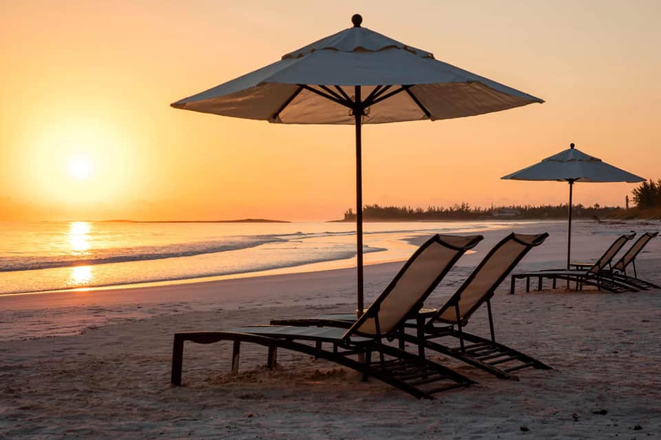 Beach sunset view with two lounge chairs under large umbrella on sandy shore at dusk