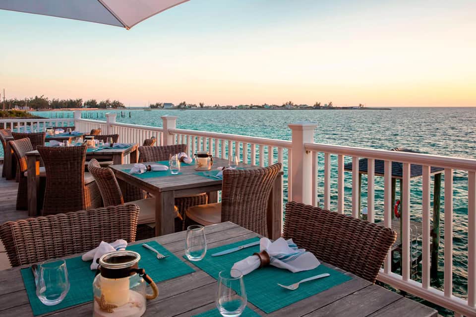 Waterfront dining deck with rattan chairs, turquoise placemats, and ocean sunset view