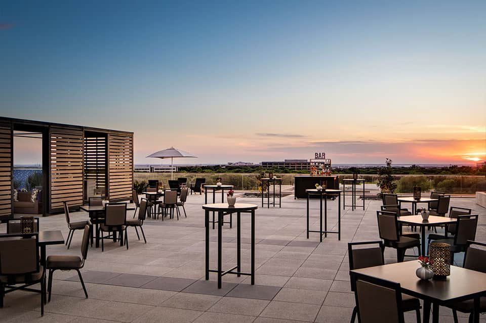 Rooftop terrace bar at sunset with lounge seating, panoramic landscape views, and modern architecture
