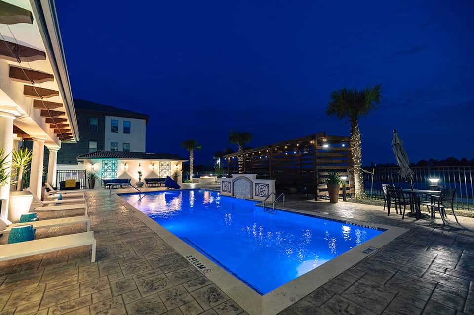 Illuminated rectangular resort pool at night with white lounge chairs and palm trees
