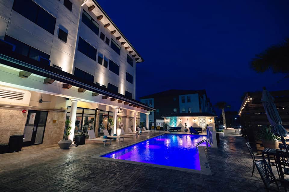 Modern resort pool illuminated at night with blue lighting, lounge chairs, and lit building facade