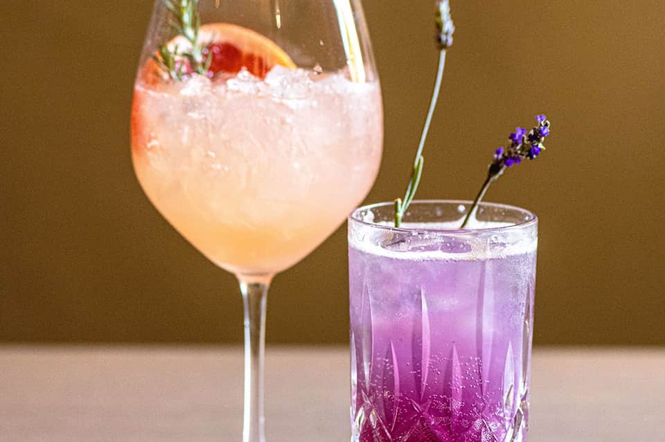 Two colorful cocktails with garnishes on neutral background, one peach one purple