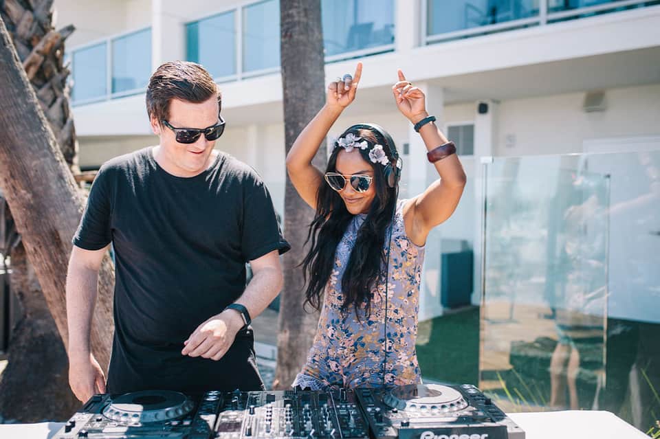 DJ performing at poolside with two people at mixing equipment near modern hotel building