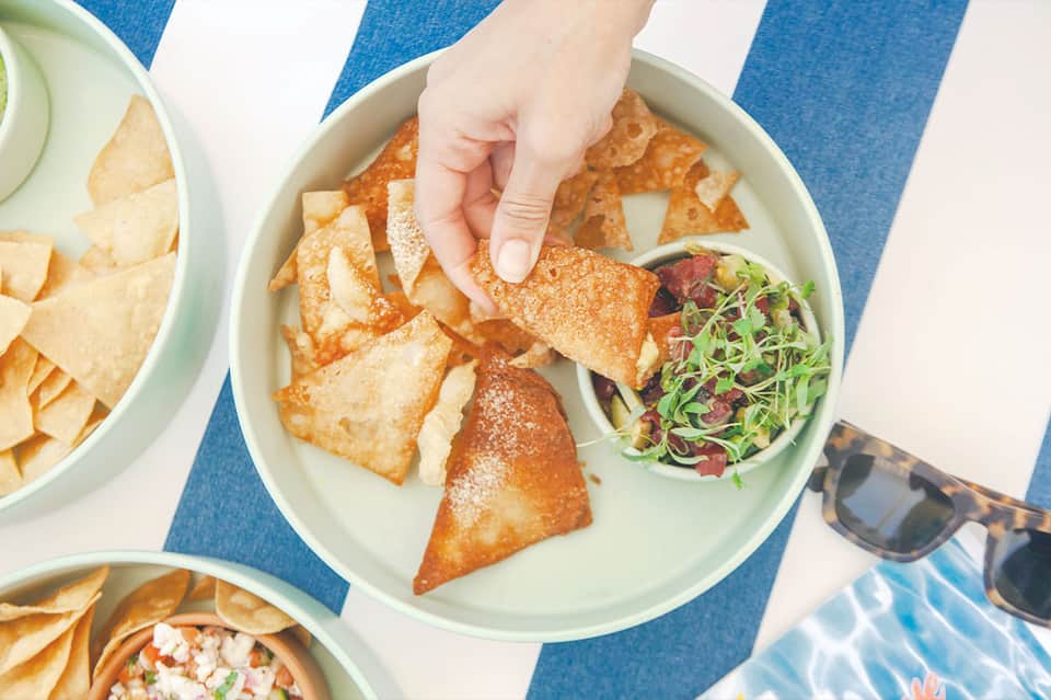 Hand holding fried wonton appetizers with salsa dip on blue and white striped beach umbrella