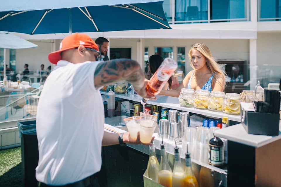 Outdoor beachfront bar with bartender serving drinks to guests under umbrella