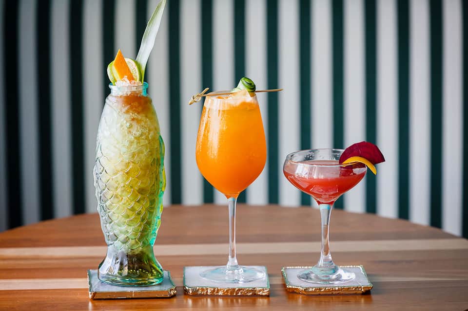 Three tropical cocktails in varied glasses with fruit garnishes on wooden table