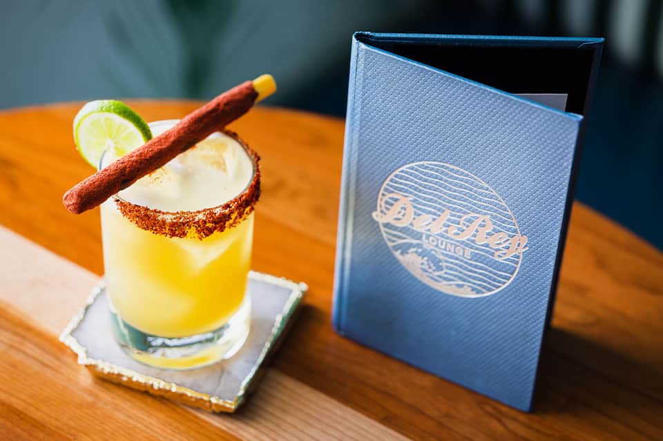 Craft cocktail with spiced rim, lime wheel, and cinnamon stick next to blue menu on wooden table