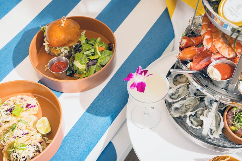 Poolside dining spread with tacos, burgers, oysters, and cocktail on striped blue and white umbrella