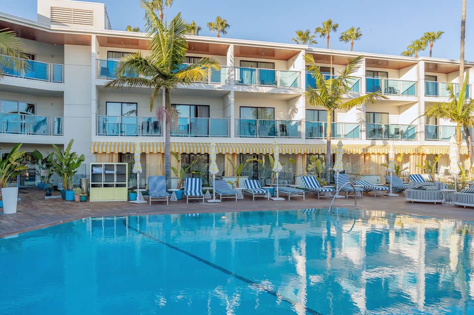 Resort swimming pool with turquoise water, white multi-story building, palm trees, and loungers