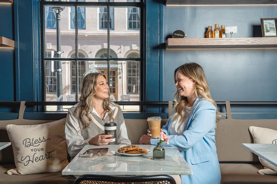 Two women chatting over coffee in a contemporary café with large windows and blue accents