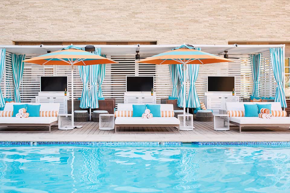 Luxury poolside lounge area with white daybeds, orange umbrellas, turquoise curtains and cabanas