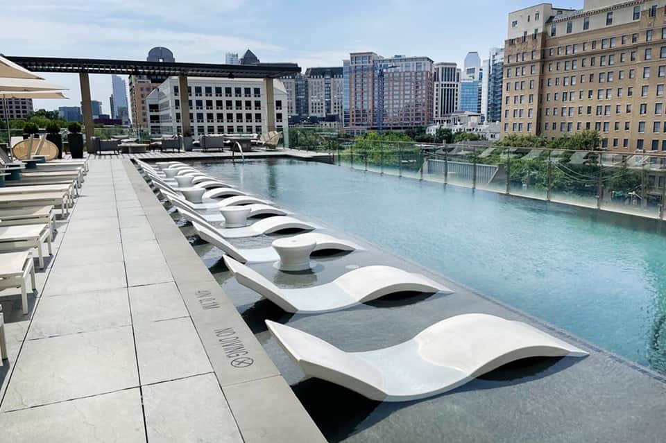 Modern white lounge chairs arranged along turquoise infinity pool with urban skyline and buildings