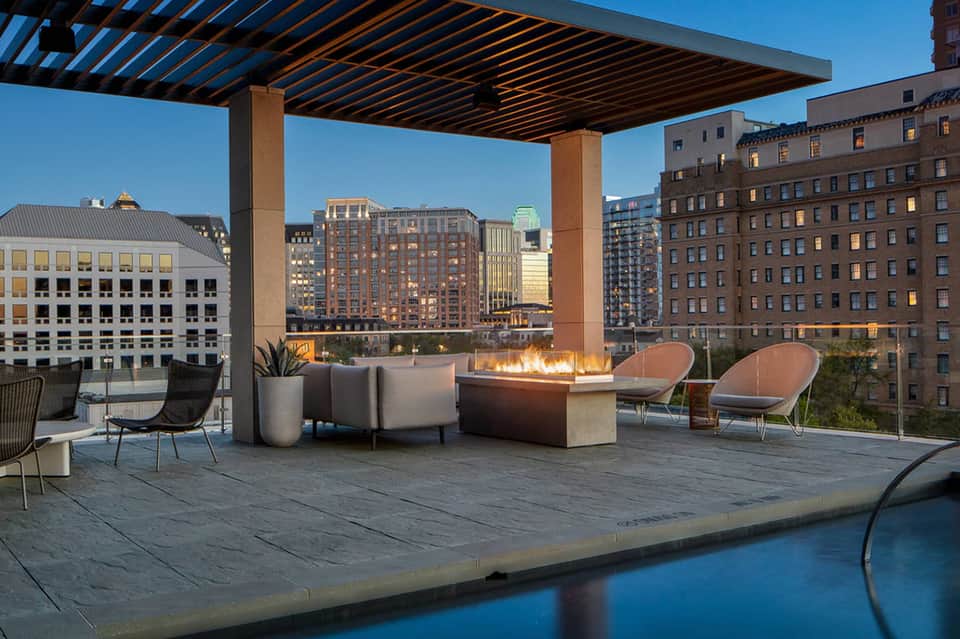Modern rooftop lounge with fire pit, contemporary seating, pergola, and city skyline views at dusk