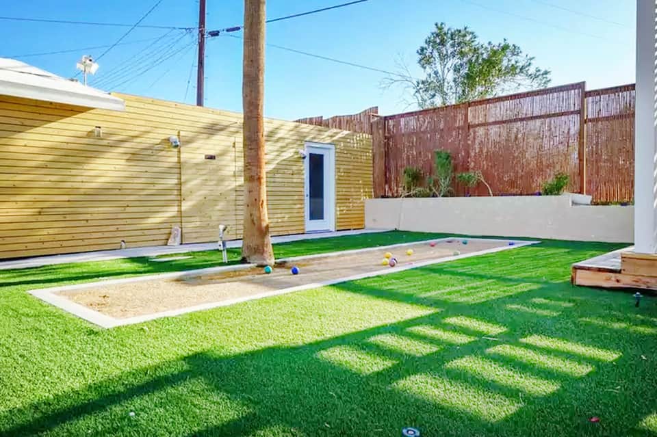 Backyard with artificial turf, sandbox, cornhole game, yellow cottage, and rusted metal fence