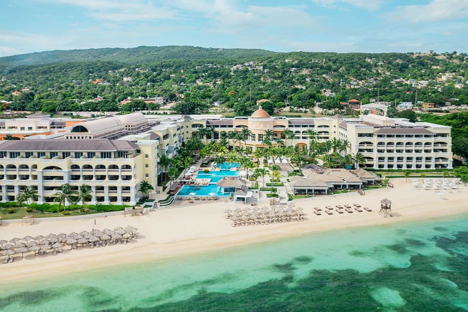 Beachfront all-inclusive resort with white buildings, turquoise pools, and lush hillside backdrop
