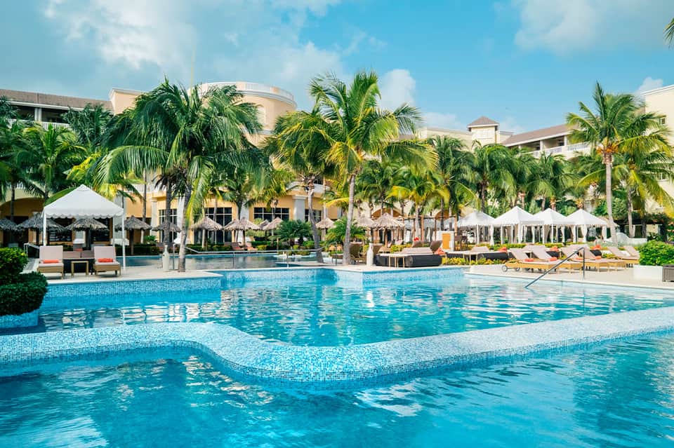 Expansive luxury resort pool with white umbrellas, palm trees, and multi-story building