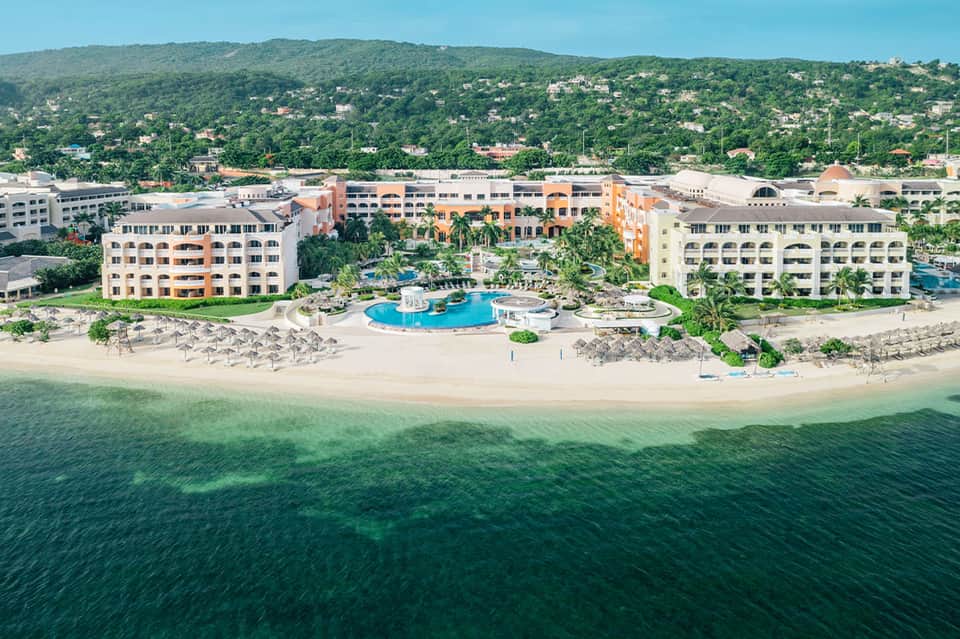 Beachfront resort aerial view showing blue pool, sandy beach with umbrellas, colorful buildings, and tropical hillside landscape