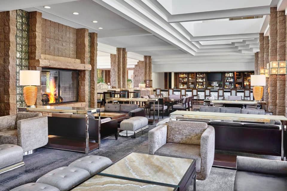 Upscale hotel lobby lounge with fireplace, stone columns, gray furnishings, and bar