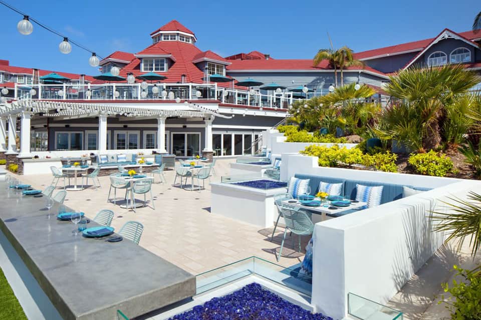 Beachfront resort terrace with blue accents, white lounge seating, and red-roofed buildings
