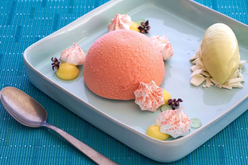 Gourmet dessert plate with pink mousse sphere, meringues, and yellow sauce on blue textured surface