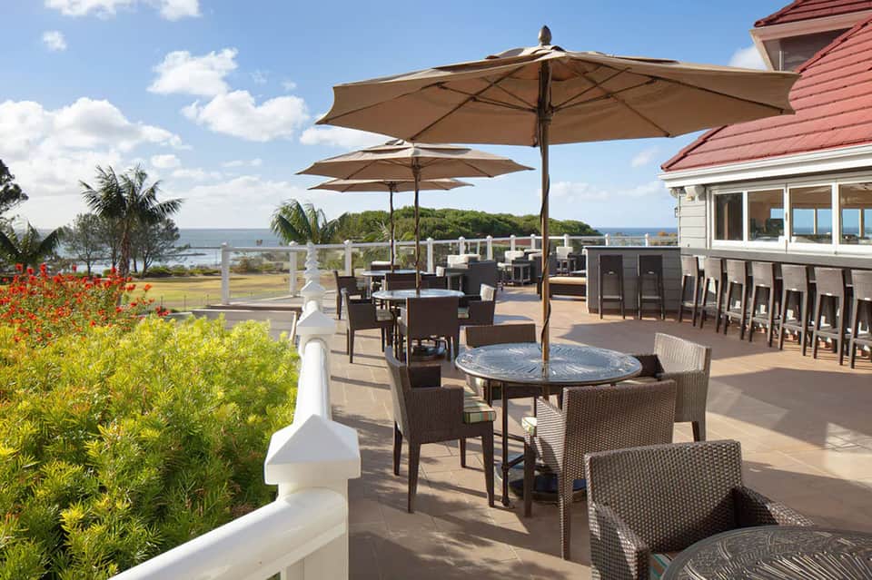 Waterfront terrace with umbrellas, wicker furniture, ocean views, and red-roofed building