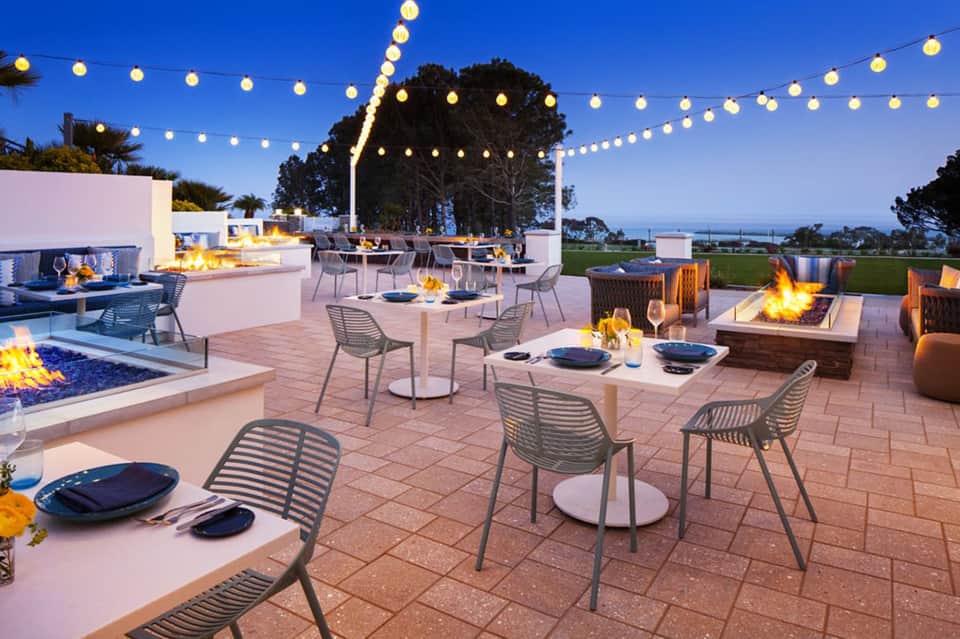 Luxury resort terrace with multiple fire pits, dining tables, string lights, fireplace features, and water views at night
