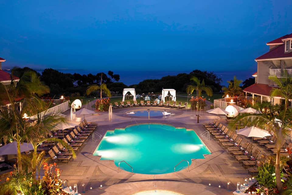 Illuminated resort pool at twilight with turquoise water, loungers, palm trees, and ocean view