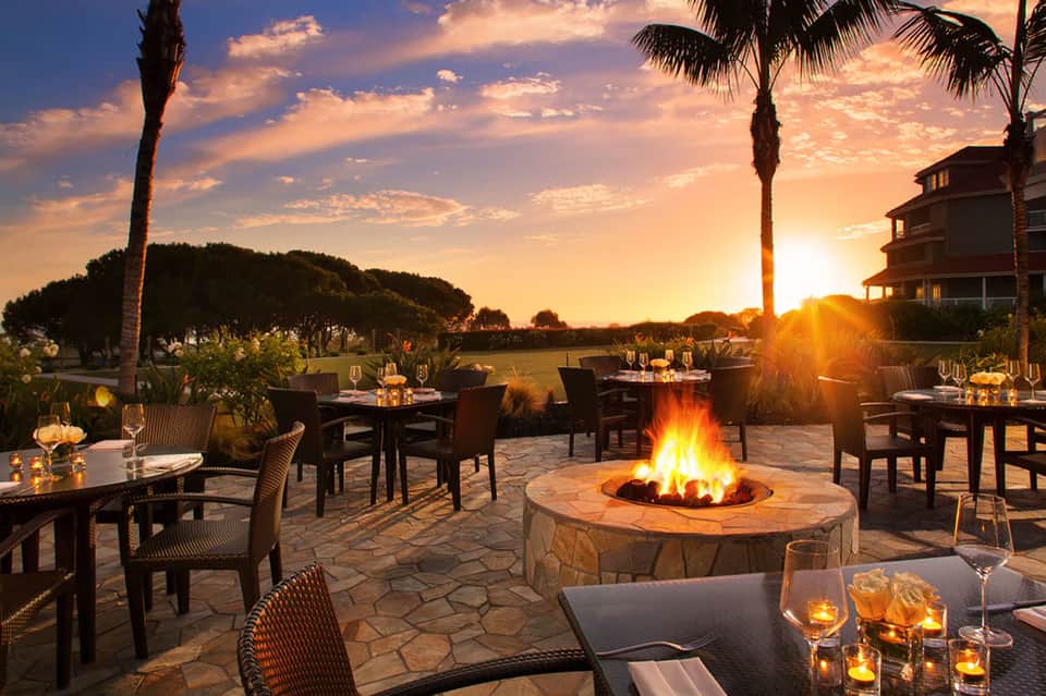 Sunset dining terrace with firepit, palm trees, restaurant tables, and golden sky backdrop