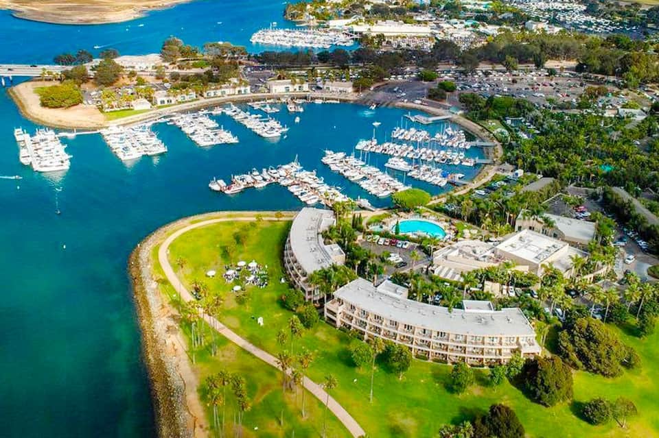 Aerial view of marina with white boats docked, curved waterfront building, and lush green grounds surrounded by blue water