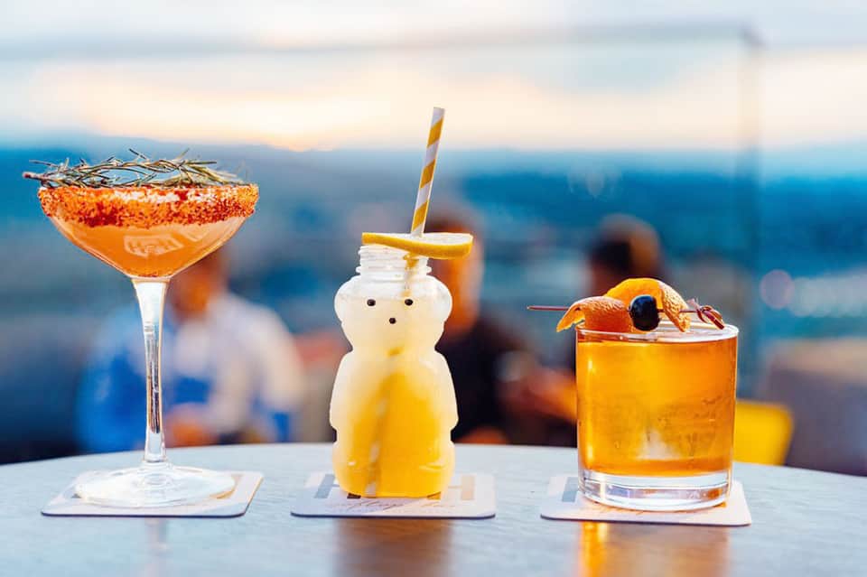Three colorful tropical cocktails with orange and citrus garnishes against an ocean backdrop