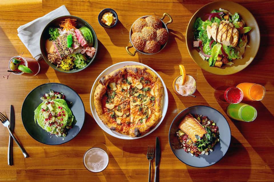 Overhead view of restaurant table with colorful dishes, pizza, salads, and fresh juices on wooden surface