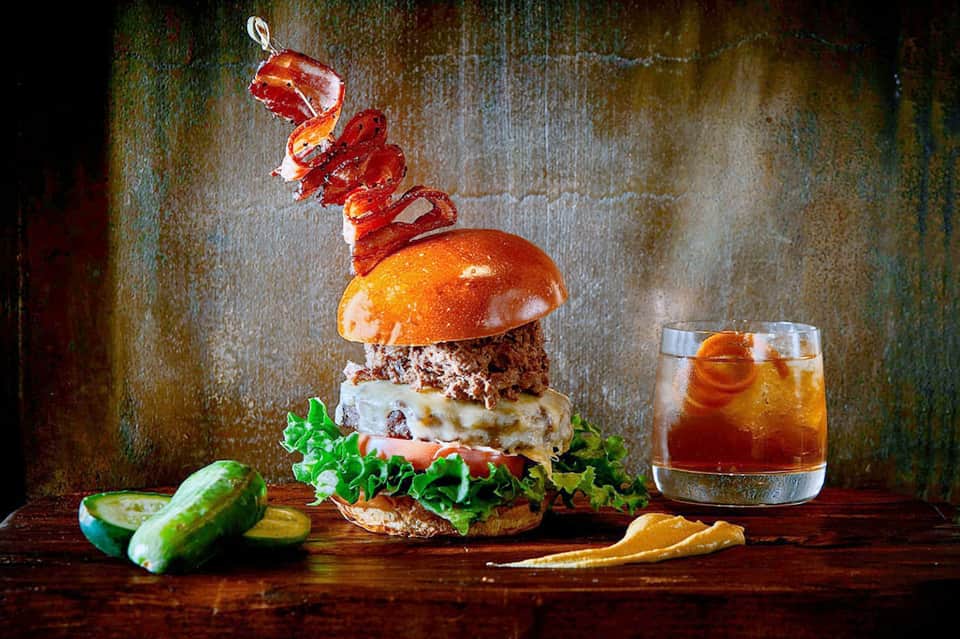 Artistic gourmet burger with red pepper garnish, served with drink and pickle sides on wooden surface