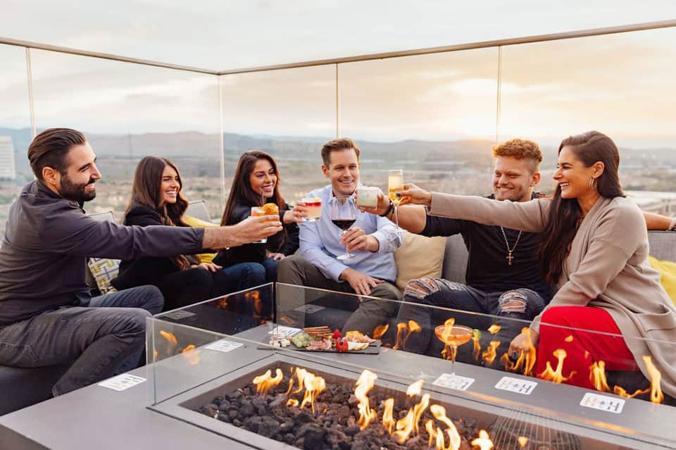 Group of six people toasting with drinks around modern fire pit lounge with panoramic valley views