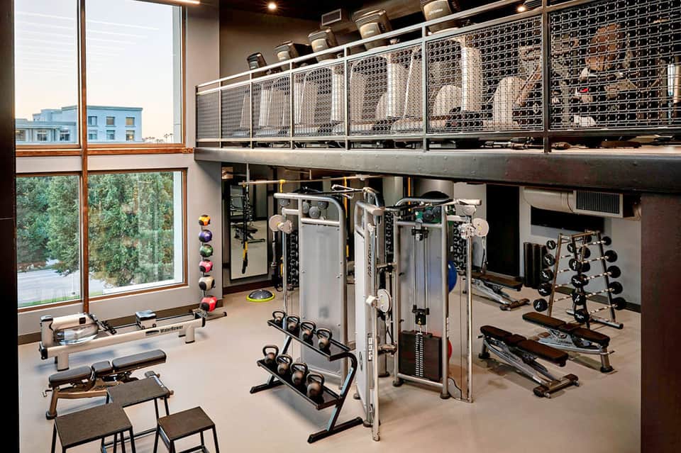 Modern hotel gym with weights, machines, large windows, and mezzanine level storage