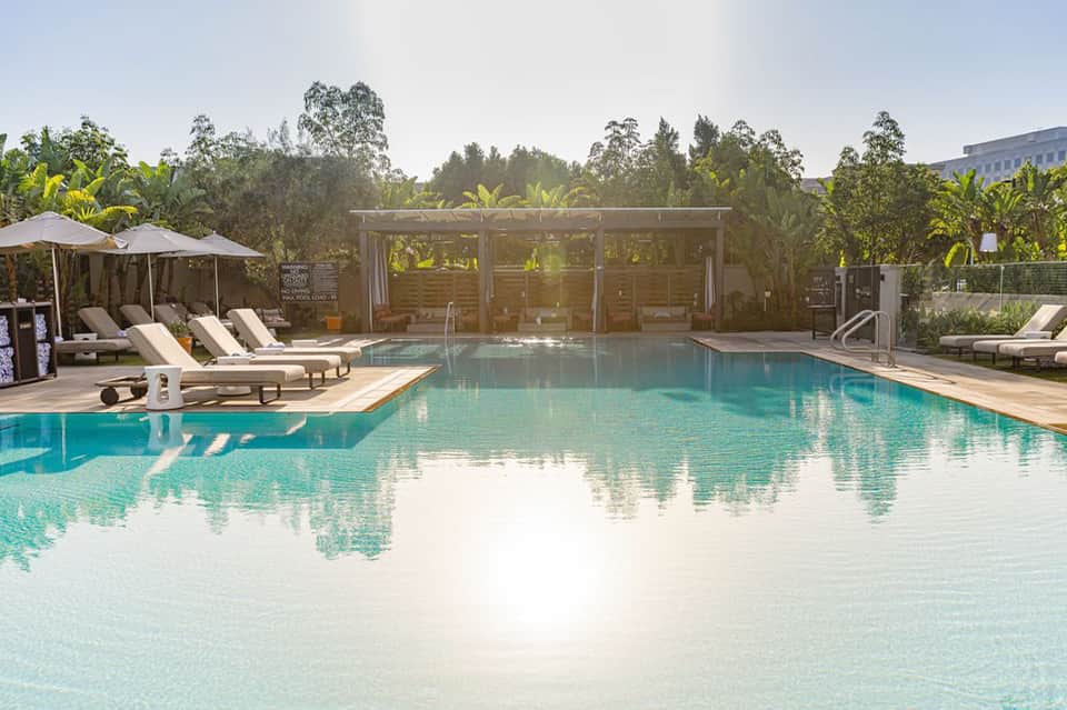 Resort pool with lounge chairs, umbrellas, modern pavilion, and surrounding vegetation