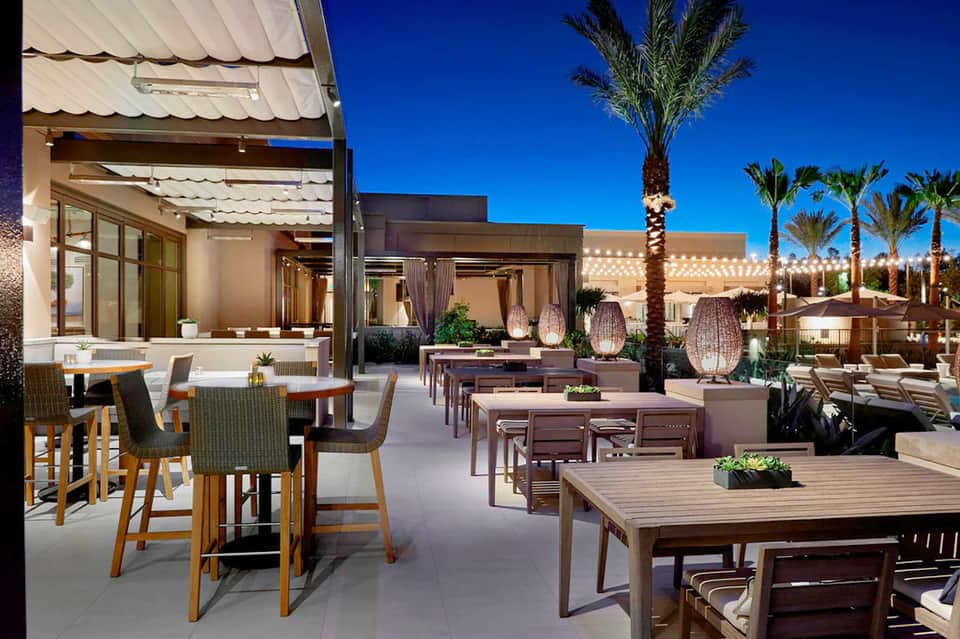Modern desert resort terrace with dining tables, bar seating, palm trees, and contemporary architectural design