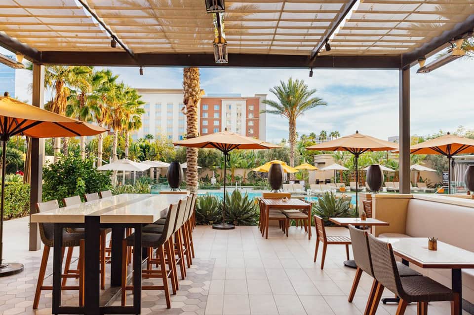 Covered poolside dining area with orange umbrellas, palm trees, and views of resort grounds
