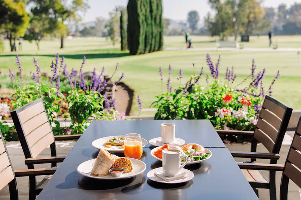 Outdoor dining table with food and drinks overlooking manicured lawn with purple flowers and cypress trees