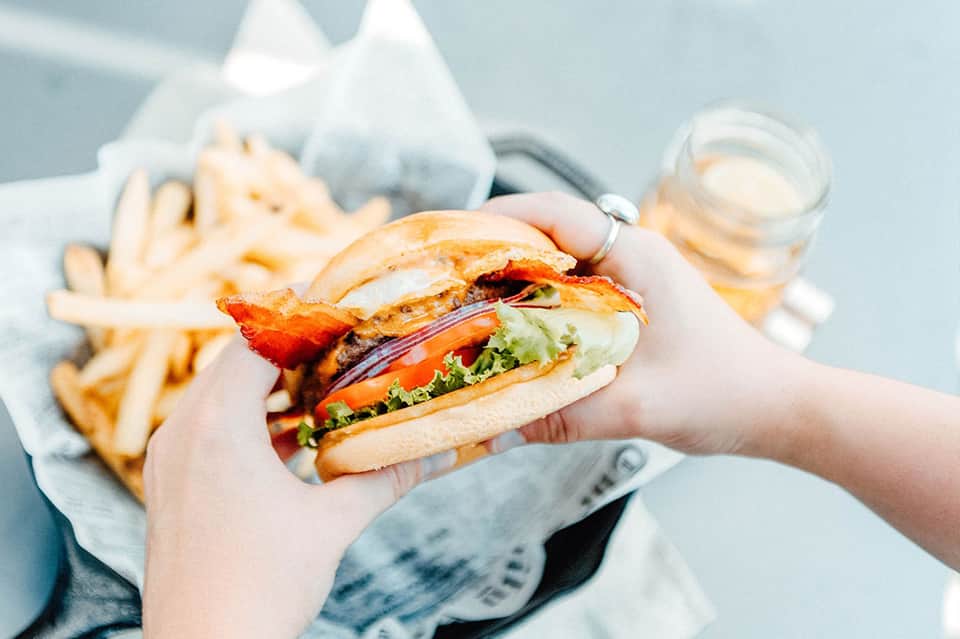 Hand holding gourmet burger with bacon and fresh vegetables next to fries