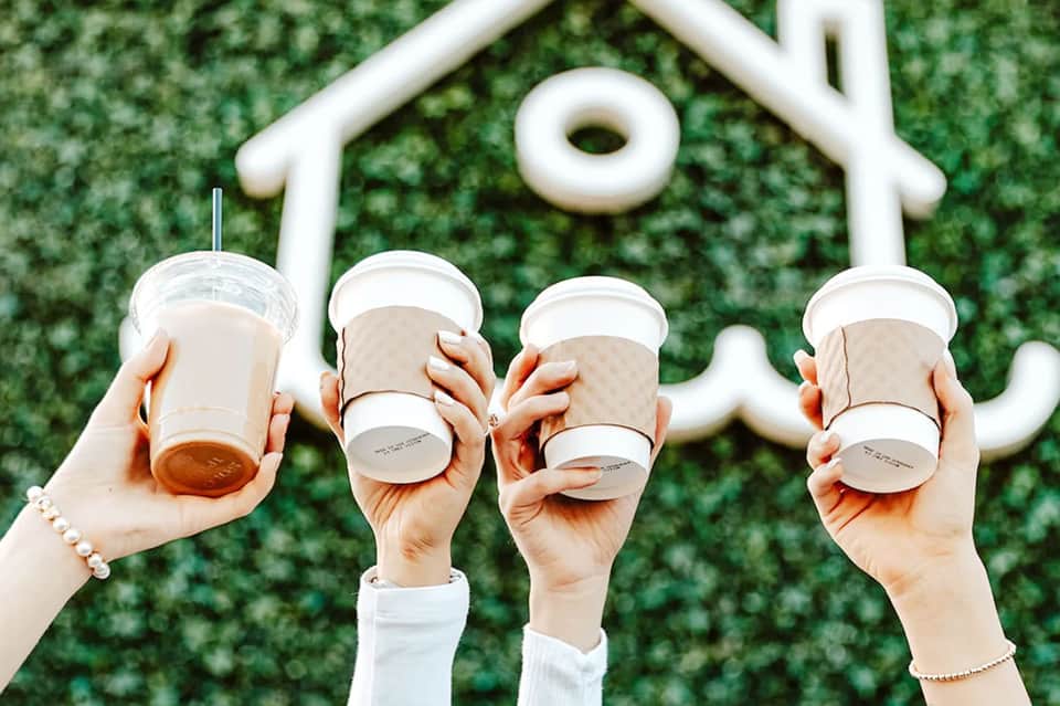 Four hands holding coffee cups in front of white decorative sign with green ivy wall backdrop