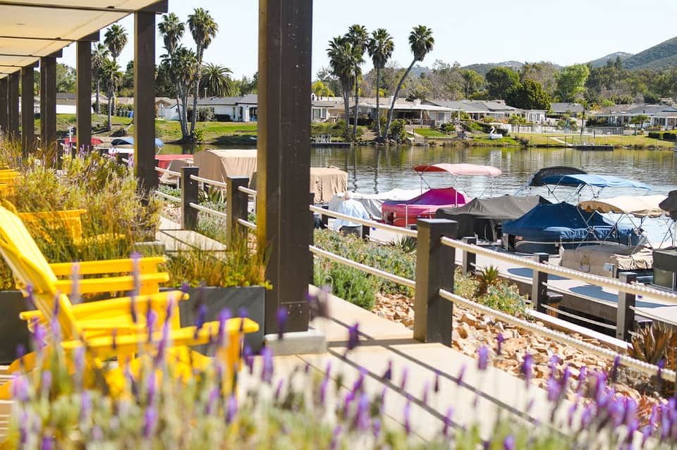 Waterfront covered patio with yellow chairs, purple flowers, boats on water, and forested hills beyond