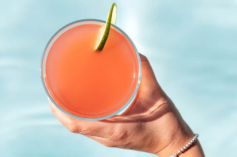 Tropical cocktail with lime garnish held against a blue sky and ocean backdrop
