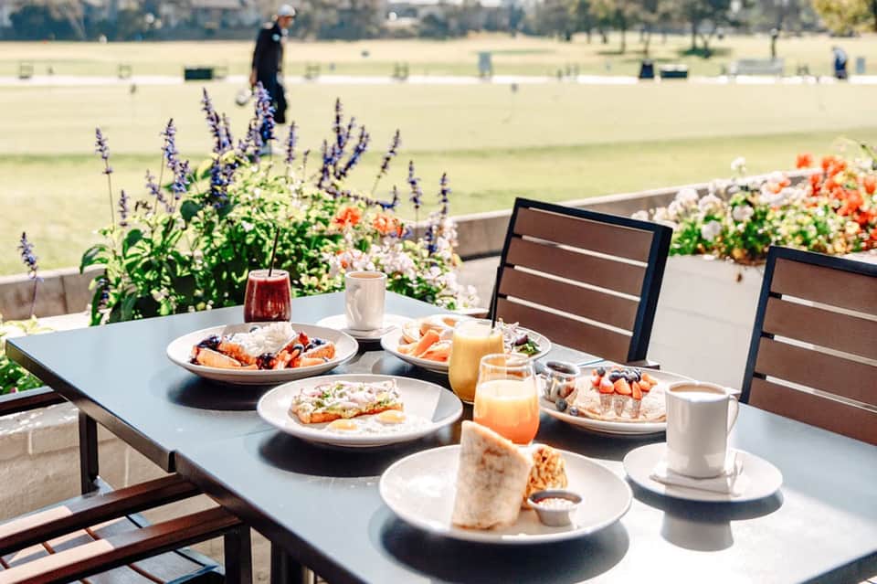 Outdoor brunch table with food and beverages, colorful flowers, overlooking a green polo field